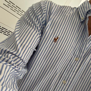 Blårandig skjorta från Polo Ralph Lauren - Snygg blårandig skjorta från Polo Ralph Lauren i custom fit. Skjortan har klassisk krage, knappar hela vägen och det ikoniska broderade logot på bröstet. Tillverkad i mjuk bomull och har långa ärmar med manschettknappar. Perfekt för en clean och fräsch look. ALDRIG använd!