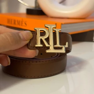Brunt skärp  - Snyggt brunt bälte med ett stort guldfärgat RLL-spänne framtill. Bältet är gjort i slätt skinn och har klassisk design med flera hål för justering. Perfekt accessoar för att lyfta din outfit med en touch av lyx.