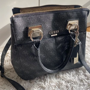 Svart handväska från Guess med gulddetaljer - Snygg svart handväska från Guess med klassiskt logomönster och guldfärgade metalldetaljer. Väskan har både handtag och justerbar axelrem, dragkedja upptill och flera fack inuti.