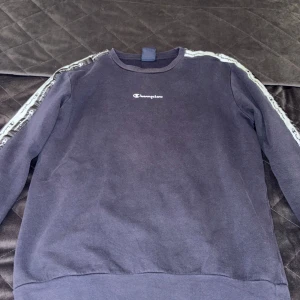 Mörkblå Champion sweatshirt - Säljer en mörkblå sweatshirt från Champion i storlek XL, passar 13/14 år Tröjan har vita band med Champion-logga längs ärmarna och broderad logga på bröstet. Skön och avslappnad passform, perfekt till chill eller streetwear. Materialet är mjuk bomull. 