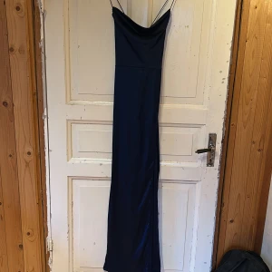 Mörkblå långklänning från NLY Eve - Supersnygg mörkblå långklänning från NLY Eve med tunna axelband som korsas i ryggen. Klänningen har en elegant draperad urringning, slits nertill och är figurnära med slät, glansig yta. 