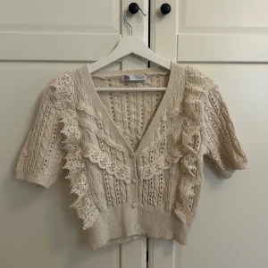 Beige stickad kofta med spets från Zara - Supersöt beige stickad kofta från Zara med korta ärmar, v-ringning och knappar framtill. Dekorerad med romantiska spetsvolanger och fina stickade detaljer. Perfekt för dig som gillar en feminin och trendig stil.