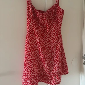 Klänning - Supersöt röd kortklänning med vita blommor och smala axelband som knyts upptill. Klänningen har en liten rosett framtill och smockad rygg för extra komfort. 