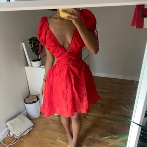 Röd omlottklänning med puffärmar - Säljer en röd kortklänning med djup v-ringning och puffiga kortärmar. Klänningen har snörning i ryggen och öppet parti baktill, vilket ger en unik och trendig look. Materialet är lätt och har en fin struktur, perfekt för dig som vill sticka ut.