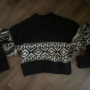 Svart stickad tröja med fransar - Säljer en svart stickad tröja med vita geometriska mönster över bröstet och ärmarna. Tröjan har långa ärmar och coola fransdetaljer längs sidorna. Ribbad hals och muddar ger en skön vibe. Perfekt för dig som gillar statement-plagg och vill sticka ut. Använd 2 gånger 