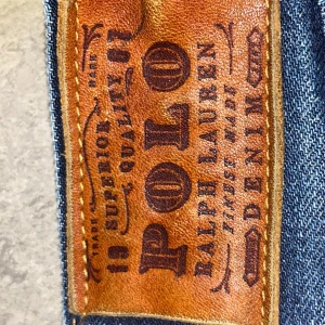 Blå jeansshorts från Polo Ralph Lauren - Snygga blå jeansshorts från Polo Ralph Lauren med råa, fransiga benslut och klassiska fem fickor. Shortsen har guldfärgade sömmar, dragkedja och en liten röd RL-brodering vid fickan. Perfekta för en chill sommarstil.