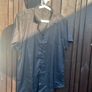 Svart kortärmad skjorta - Stilren svart skjorta med korta ärmar och klassisk krage. Skjortan har knäppning framtill och är tillverkad i ett slätt, lätt material som ger en snygg och enkel look. Perfekt för dig som gillar minimalistisk stil.