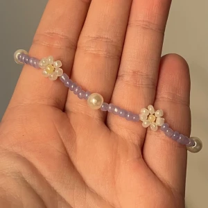 Lila pärlarmband med blommor - Jag gör egna smycken. Supergulligt handgjort armband med små lila pärlor och vita blommor, plus några större pärlor som detaljer. Armbandet har justerbar kedja i silverfärg och är perfekt för att ge din outfit en söt och lekfull touch.