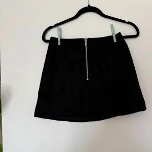 Svart kortkjol med dragkedja - Snygg svart kortkjol med rak passform och en silvrig dragkedja framtill. Kjolen är enkel och stilren, perfekt att matcha med en cool t-shirt eller stickad tröja. Materialet känns mjukt och har en lätt struktur.