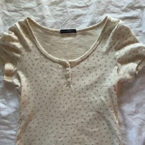 Topp från Brandy Melville - Supersöt beige topp från Brandy Melville med små blommor, korta puffärmar och rundad halsringning. Toppen har knappar framtill och är i mjuk bomull, perfekt för en chill och söt look.