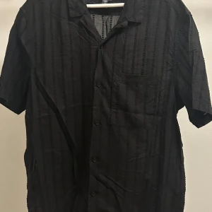 Svart randig skjorta från H&M - Svart skjorta från H&M i regular fit med diskreta vertikala ränder och kort ärm. Skjortan har klassisk krage, knäppning framtill och en bröstficka. 