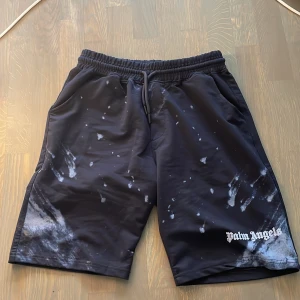 Palm Angels svarta shorts med tryck - Svarta shorts från Palm Angels i 100% bomull med vit logga på benet och coolt stänkmönster i vitt. De har elastisk midja med snörning och dragkedja i fickorna. Perfekta för dig som gillar streetwear och vill sticka ut lite extra.