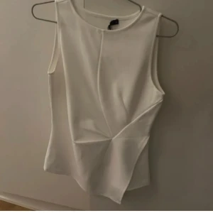 Vit ärmlös topp från Bikbok - Säljer en stilren vit ärmlös topp från Bikbok i storlek S. Toppen har en asymmetrisk skärning och coola veckdetaljer framtill som ger en modern look. Perfekt att matcha med jeans eller kjol för en clean och trendig outfit.
