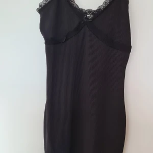 Svart ribbad klänning från H&M Divided - Svart ribbad kortklänning från H&M Divided med spetsdetaljer längs urringningen och en liten rosett framtill. Ärmlös modell med smala axelband och v-ringad hals. Perfekt för dig som gillar en enkel men ändå söt stil.