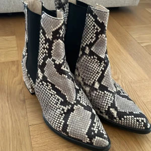 Chelsea boots med ormskinnsmönster - Snygga chelsea boots med spetsig tå och chunky klack i ormskinnsmönstrat syntetmaterial. Skorna har svart resår på sidorna och dragflik bak för enkel påtagning. Färgerna går i beige, svart och grått för en edgy look.