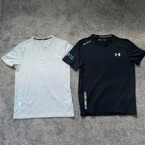 Under Armour t-shirts i svart och grå - Två snygga Under Armour t-shirts, en i svart och en i ljusgrå. Båda har korta ärmar, rund hals och coola loggor samt textdetaljer i regnbågsfärger och svart. Perfekta för träning eller chill, tillverkade i mjukt och lätt polyester-material.