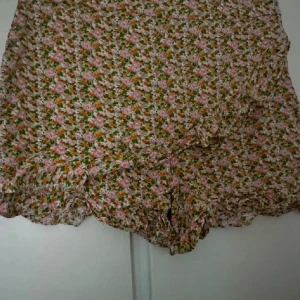 Blommiga volangshorts i pastell - Supersöta shorts med blommigt mönster i rosa, orange och grönt. Lätta och luftiga med volangdetaljer och omlottkänsla. Perfekta för varma dagar när du vill ha något både bekvämt och trendigt.