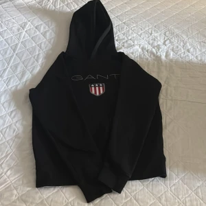 Svart hoodie från GANT med logga - Svart hoodie från GANT med broderad logga och sköld på bröstet. Klassisk modell med huva och långa ärmar, perfekt för en chill och stilren look. Mjuk insida och ribbade muddar för extra komfort.