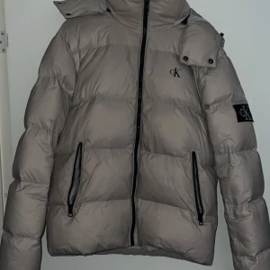 Beige pufferjacka från Calvin Klein - Säljer en beige pufferjacka från Calvin Klein Jeans med svart CK-logga på bröstet och ärmen. Jackan har huva, två dragkedjefickor och hög krage. Perfekt för kalla dagar och riktigt snygg till streetwear-stilen.