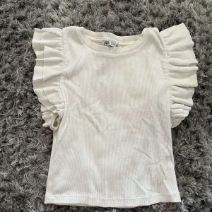 Vit volangtopp från Zara - Supersöt vit topp från Zara med breda volanger längs axlarna. Toppen är ärmlös och har en rund halsringning. Perfekt för dig som gillar en feminin och trendig stil. Materialet är mjukt och lätt ribbat för extra komfort.