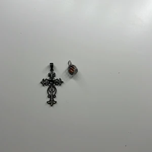 Två coola berlocker - Säljer två snygga hängen: ett korsformat hänge i mörk silverfärg med glittrande stenar och detaljerad design, samt ett runt hänge med en orange sten inuti en spiral av silverfärgad metall. Perfekta att mixa och matcha till kedja.