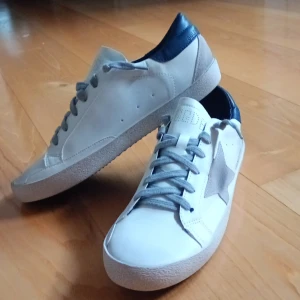Golden Goose vita sneakers med stjärna - Golden Goose sneakers i vitt skinn med grå mockadetaljer och en ikonisk stjärna på sidan.  Skorna är storlek 44✨ Skorna har grå snörning, blå hälkappa och platt sula. Kommer med original dustbag. Perfekta för dig som gillar en clean men ändå unik look.