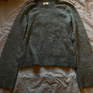 Grå stickad tröja med bred mudd - Mysig grå stickad tröja med rund halsringning och bred ribbad mudd vid ärmslut och nederkant. Perfekt för lager-på-lager och enkel att matcha med jeans eller kjol. Klassisk modell med lång ärm och mjuk känsla.