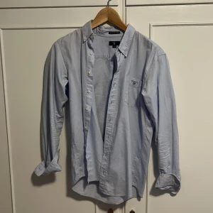 Ljusblå skjorta från GANT, 170 cm - Snygg ljusblå skjorta från GANT med klassisk button down-krage och bröstficka med broderad logga. Skjortan är i bomull och har långa ärmar samt knappar framtill. Perfekt för en clean och stilren look.