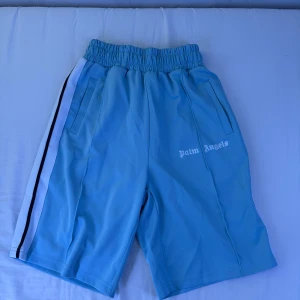 Blå Palm Angels shorts; (oanvända)  - (Pris kan diskuteras vid snabb affär) Snygga blå shorts från Palm Angels med vit och svart rand längs sidorna. Modellen har elastisk midja och två fickor framtill. Palm Angels-logga tryckt på ena benet. Perfekta för sommaren och streetwear-stil.