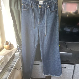 Baggy jeans Ganni - Snygga blå jeans från Ganni med breda ben och råa kanter nertill. Klassisk femficksmodell med snörning i midjan och Ganni-logga på bakfickan. Jeansen har en avslappnad passform och är tillverkade i kraftig denim. Storlek 29/M