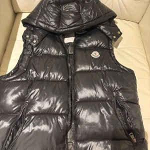 Moncler väst - Snygg svart dunväst från Moncler med glansig finish och avtagbar huva. Västen har dragkedja framtill, två fickor med dragkedja och klassisk Moncler-logga på bröstet.