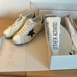 Golden Goose vita sneakers med svart stjärna - Golden Goose sneakers i vitt skinn med svart stjärna på sidan och mörkblå hälkappa. Klassisk låg modell med snörning och platt sula. Snyggt slitna detaljer och retrovibe, perfekt för dig som gillar streetstyle och exklusiva märken. Har kvitto!