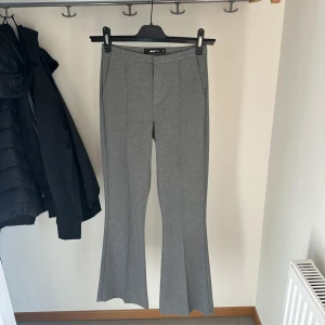 Grå bootcut kostymbyxor från Gina Tricot - Snygga grå kostymbyxor från Gina Tricot med bootcut-ben och pressveck framtill. Byxorna har en klassisk midja och är perfekta för en clean och stilren look. Materialet är mjukt och ger en dressad känsla.