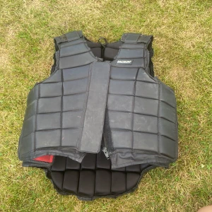 Svart skyddsväst Jacson Junior XL - Skyddsväst från Jacson i svart med rutmönstrad vaddering. Västen har justerbara axelband, breda sidospännen och dragkedja framtill. Perfekt för ridning och ger bra skydd över bröst och rygg. Tillverkad i slitstarkt syntetmaterial.   Dammig lite repor men funkar fint. 