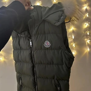 Olivgrön dunväst från Moncler - Snygg olivgrön dunväst från Moncler med pösig huva och fluffig pälsdetalj. Västen har två dragkedjefickor, quiltad design och klassisk Moncler-logga på bröstet. Perfekt för lager-på-lager och riktigt street. Dragkedja framtill och cool passform.