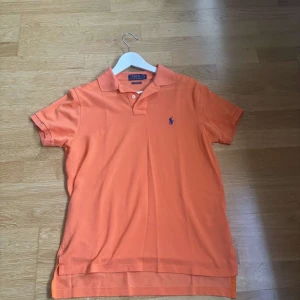 Orange pikétröja från Polo Ralph Lauren - Säljer en orange pikétröja från Polo Ralph Lauren med klassisk krage och korta ärmar. Tröjan har den ikoniska blå loggan broderad på bröstet och är tillverkad i mjuk bomull. Perfekt för en avslappnad och stilren look.