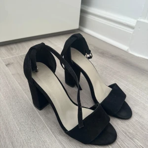 Svarta klackskor NELLY storlek 37 - Stilrena svarta sandaler med bred klack och öppen tå, klacken är 10 cm.. Skorna har en enkel rem över tårna och en smal rem runt vristen med spänne. Tillverkade i mockaliknande material som ger en lyxig känsla. Perfekta för dig som gillar minimalistisk och trendig stil. 