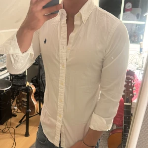 Vit skjorta från Ralph Lauren - Klassisk vit skjorta från Ralph Lauren med mörkblå broderad logga på bröstet. Skjortan har lång ärm, knappar hela vägen och en snygg, slim passform. Tillverkad i bomull och passar perfekt till jeans eller chinos.
