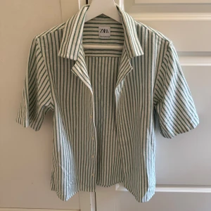 Randig skjorta från Zara - Kortärmad skjorta från Zara i vit och grön randig design. Skjortan har klassisk krage, knäppning framtill och är tillverkad i mjuk bomull. Använd ett fåtal gånger men har inga tecken på användning alls 