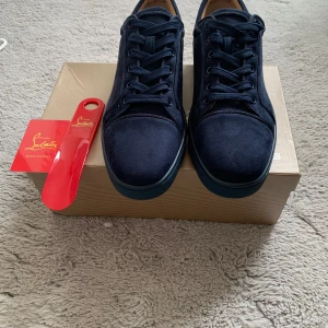 Mörkblå sneakers från Christian Louboutin - Exklusiva mörkblå sneakers i mocka från Christian Louboutin med klassisk röd sula och broderad logga på hälen. Skorna har rund tå, snörning och stilren design som sticker ut med den ikoniska röda undersidan.