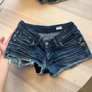 G-Star Raw jeansshorts med snygga detaljer - Snygga mörkblå jeansshorts från G-Star Raw med råa, fransiga kanter och slitna detaljer. Klassisk femficksmodell med låg midja och coola kontrastsömmar. Perfekta för sommardagar när du vill ha en avslappnad och edgy look.