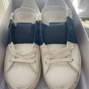 Valentino vita/blå sneakers - Snygga sneakers i vitt skinn med bred blå panel över mitten. Klassisk rund tå, vita snören och coola nitar på sulan. Perfekta för dig som vill ha en stilren men ändå unik look. Storlek 46 men passar mer för de som är 45 ish