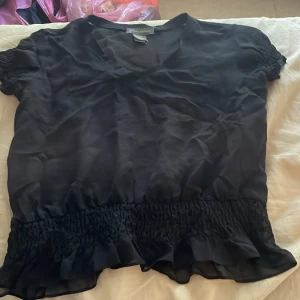Svart silkesblus från Banana Republic - Supersnygg svart blus i 100% silke från Banana Republic. Modellen har v-ringning, korta ärmar med smock och volangdetaljer samt bred smockad midja med volangkant. Tunn och lyxig känsla, perfekt för dig som gillar stilrena och feminina plagg.