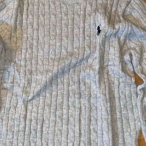 Grå kabelstickad tröja Polo Ralph Lauren - Kabelstickad grå tröja från Polo Ralph Lauren med klassisk rund halsringning och broderad logga på bröstet. Tröjan är i mjuk ullblandning och har lång ärm. Perfekt för dig som gillar stilren och tidlös design.