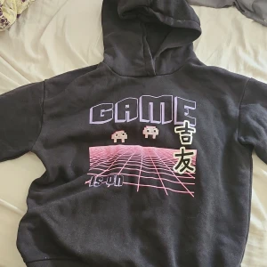 Svart hoodie med gaming-tryck från Lindex - Svart hoodie från Lindex med retro gaming-tryck i lila och rosa framtill, japanska tecken och texten 'LEVEL UP' på ärmen. Mjuk insida, huva och lång ärm. Perfekt för dig som älskar gaming och coola prints.
