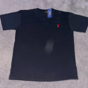 Svart t-shirt från Polo Ralph Lauren - Svart t-shirt från Polo Ralph Lauren med klassisk röd broderad logga på bröstet. T-shirten har rund halsringning och korta ärmar. Tillverkad i mjuk bomull för en skön känsla och enkel stil.
