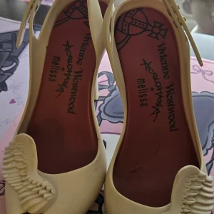 Vivienne Westwood x Melissa pumps med vingar - Unika pumps i vit färg från Vivienne Westwood x Melissa, tillverkade i syntetmaterial. Skorna har öppen tå, hög klack och en cool 3D-detalj med vingar framtill. Perfekta för dig som vill sticka ut med en edgy och lekfull stil. Fint skick förutom lite smutsiga.
