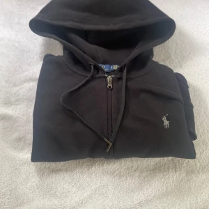Svart hoodie från Polo Ralph Lauren - Svart hoodie från Polo Ralph Lauren med dragkedja och huva. Klassisk design med broderad logga på bröstet och snörning i huvan. Tillverkad i mjuk bomull. Fickor framtil. Pris kan diskuteras.