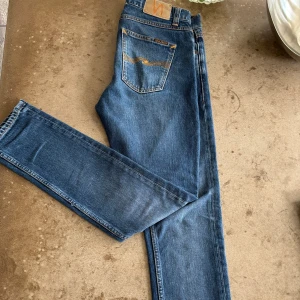 Blå jeans från Nudie Jeans, W29 L32 - Säljer ett par klassiska blå jeans från Nudie Jeans med snygga orange sömmar och ikonisk broderad bakficka. Modellen har normal passform och raka ben. Tillverkade i slitstarkt bomullsjeans, perfekta till sneakers eller boots.