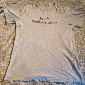 Grå Peak Performance t-shirt - Snygg ljusgrå t-shirt från Peak Performance med svart tryck på bröstet. Klassisk rund halsringning och korta ärmar. Tillverkad i mjuk bomull, perfekt för en chill och stilren look. Storleksettiket är tyvärr bortklippt men tvättrådslappen är kvar, annars i bra skick. Är en junior tröja, storlek är tyvärr okänd men skulle säga Small.